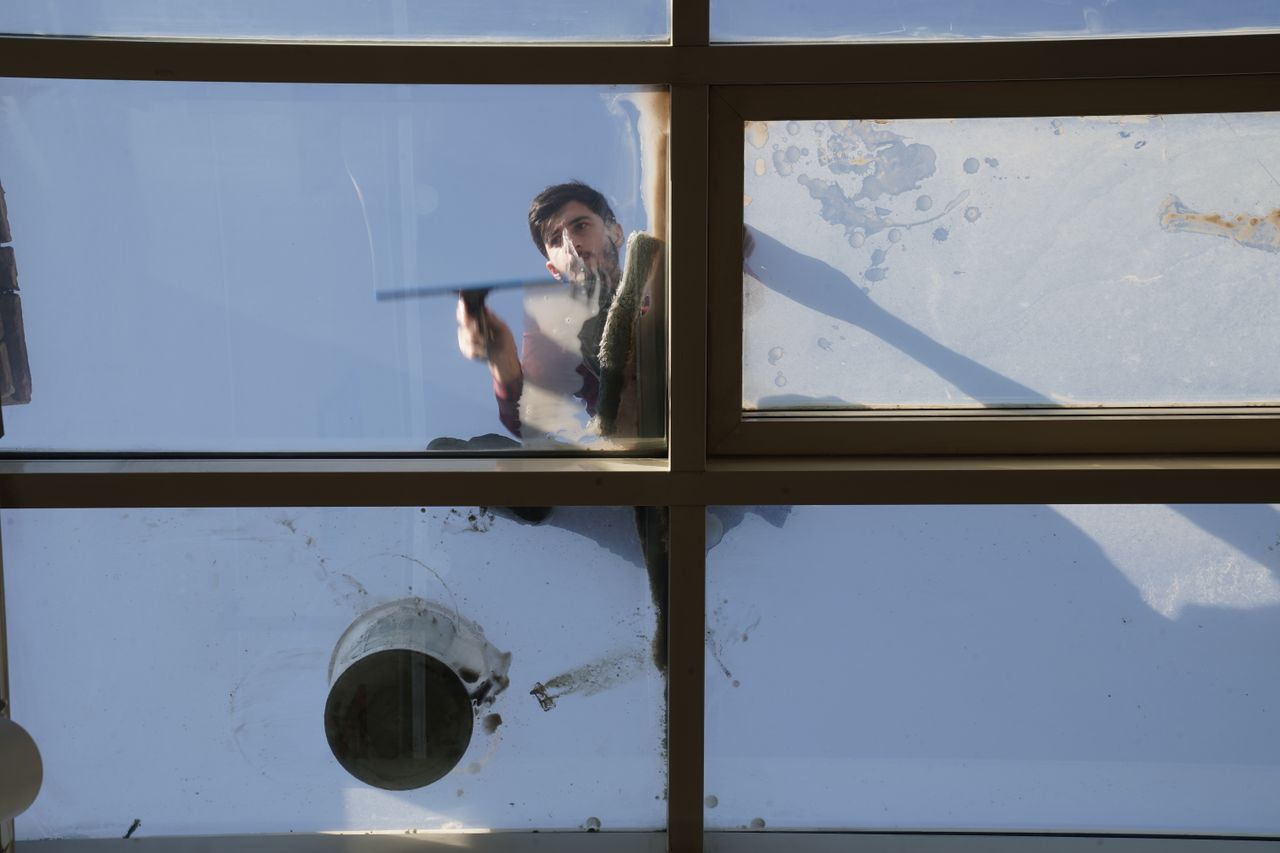 Un laveur de vitres masculin nettoie, en plein jour, la toiture vitrée extérieure depuis le dessus à l’aide d’un outil.